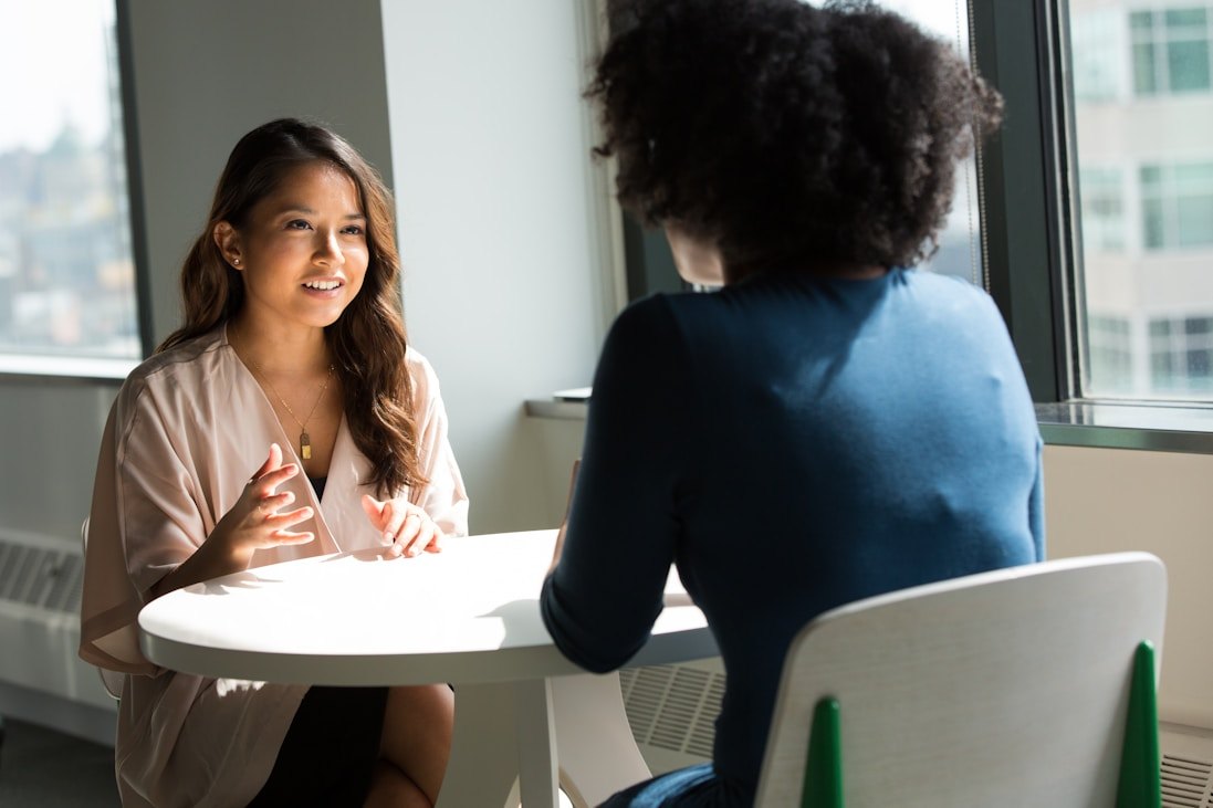 Two women in conversation across a table beside a window.